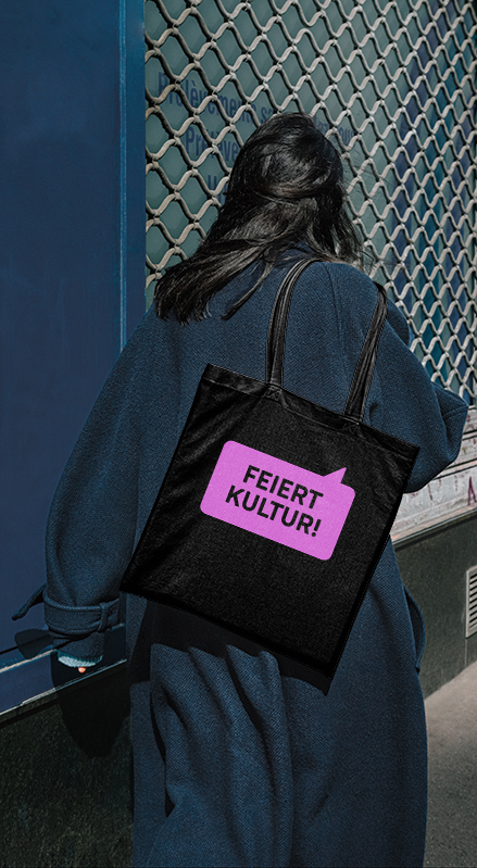 Person mit schwarzer Stofftasche der Gasteig-Kampagne „Feiert Kultur!“ vor urbaner Kulisse in München – Give-away der Jubiläumskampagne zum 40-jährigen Bestehen des Kulturzentrums.