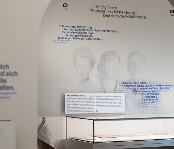 Der Blick in die Ausstellung zeigt die Perspektive durch einen gewälbten Torbogen, hinter dem man direkt auf den Wand gedruckte Personen, Zitate, eine Vitrine und Texttafeln erkennt.