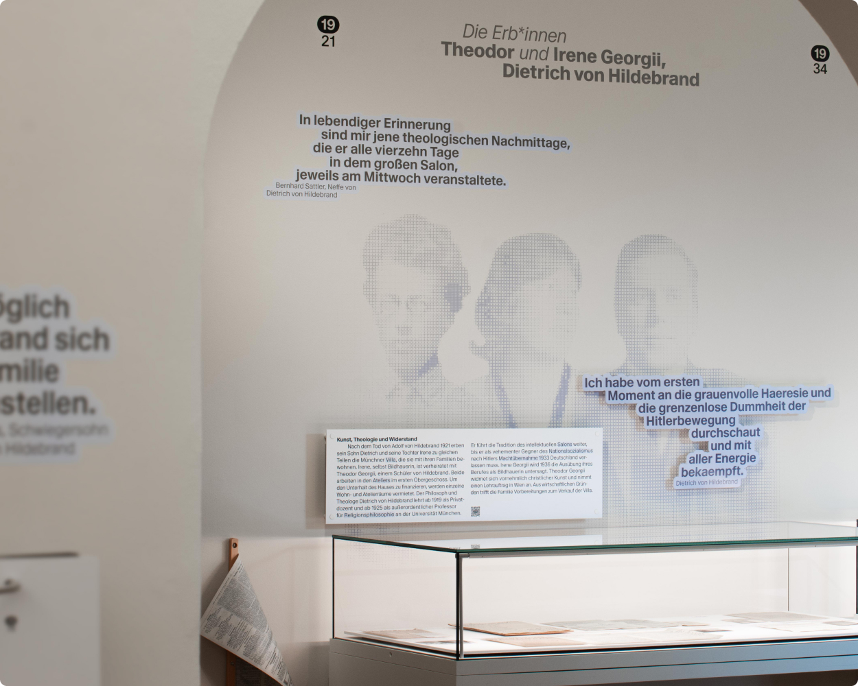 Der Blick in die Ausstellung zeigt die Perspektive durch einen gewälbten Torbogen, hinter dem man direkt auf den Wand gedruckte Personen, Zitate, eine Vitrine und Texttafeln erkennt.
