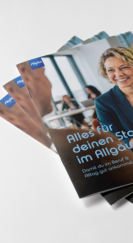 Mehrere Exemplare der CheckIn → Allgäu Broschüre 'Alles für deinen Start im Allgäu' fächerförmig aufgestellt, Cover mit lächelnder Frau im Büroumfeld – Print-Kollateral von Büro Alba