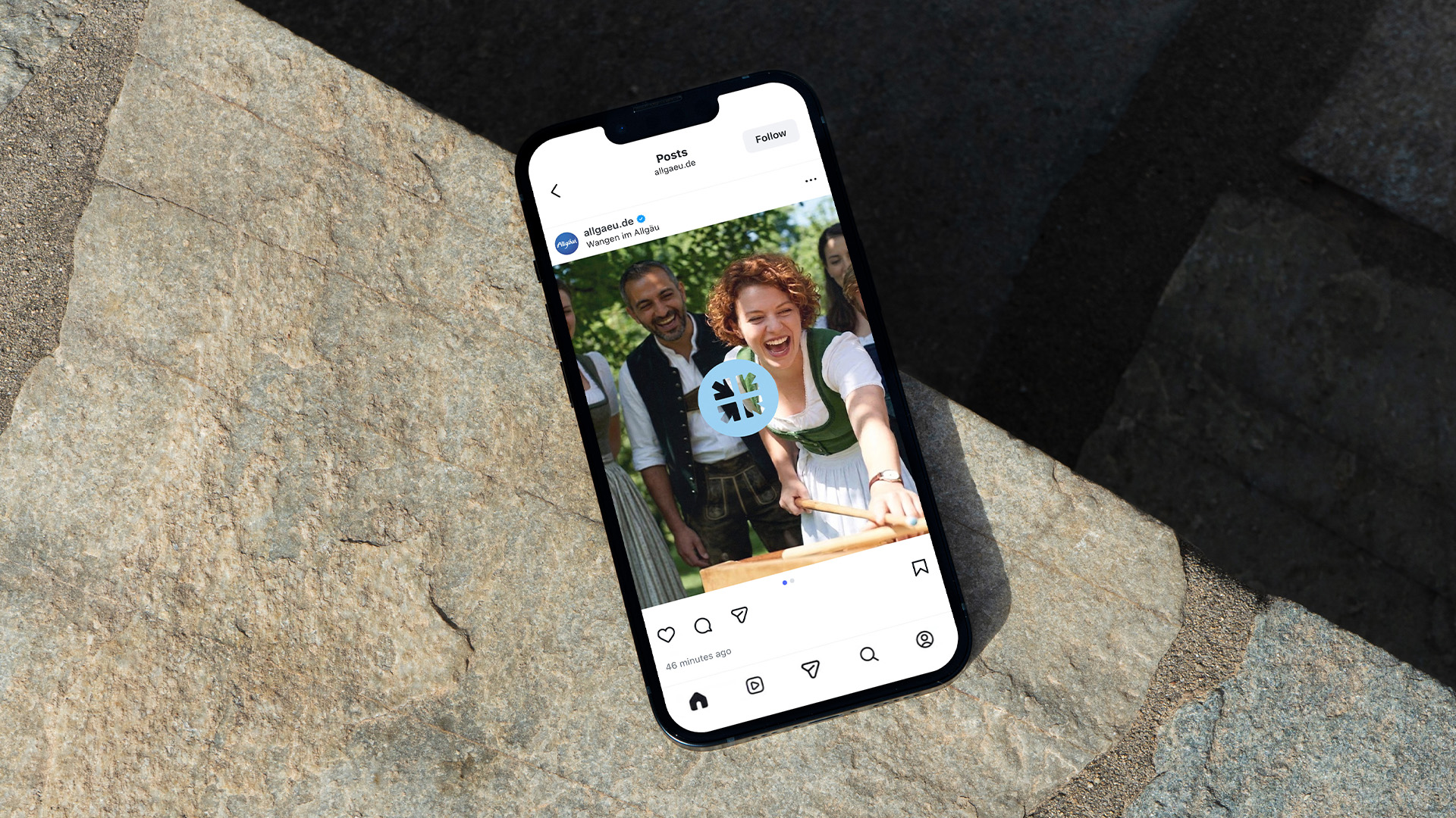 Smartphone auf einem Fels zeigt einen @allgaeu.ankommen Instagram-Post mit zwei Personen in traditioneller Allgäuer Tracht,
