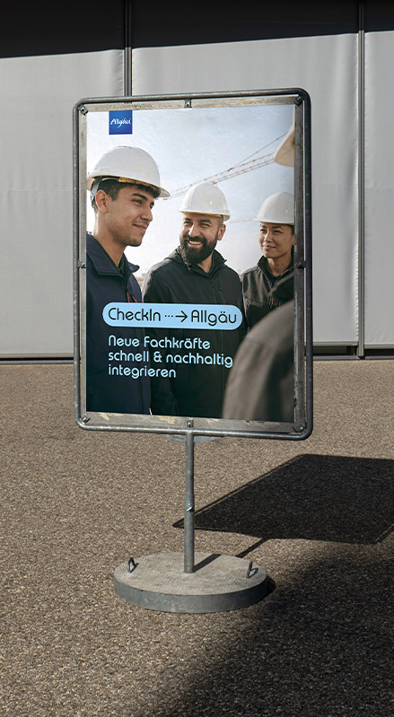CheckIn → Allgäu Poster auf freistehendem Ständer im Büroumfeld, Headline zur erfolgreichen Integration neuer Fachkräfte in Beruf und Alltag