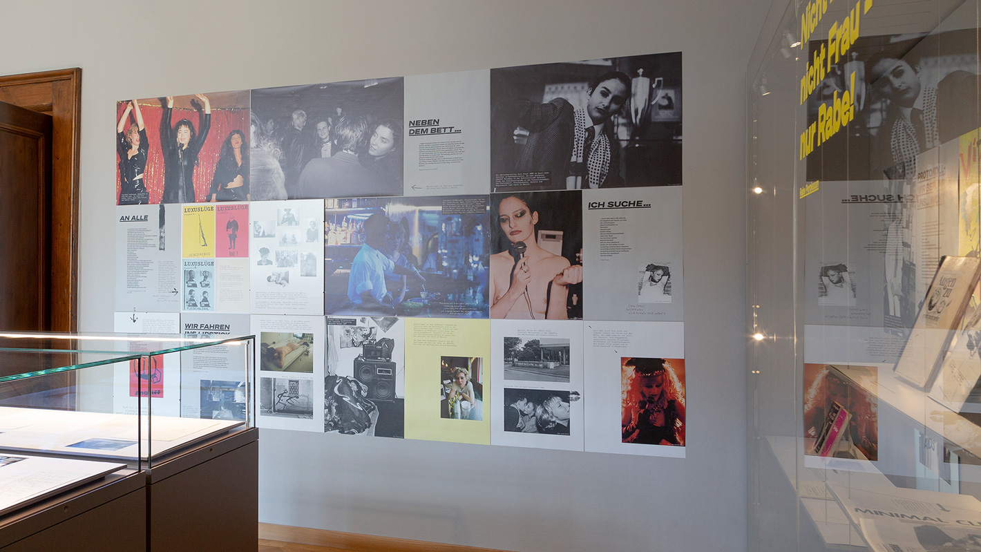 Verschiedene Fotografien, Plakate und weitere Exponate angeordnet an einer Wand der Ausstellung.