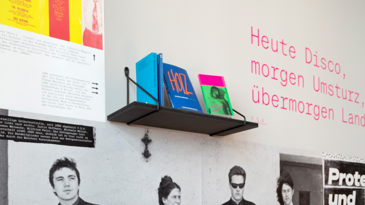 Bücherregal an der Wand der Ausstellung umgeben von Fotografien und Plakaten.