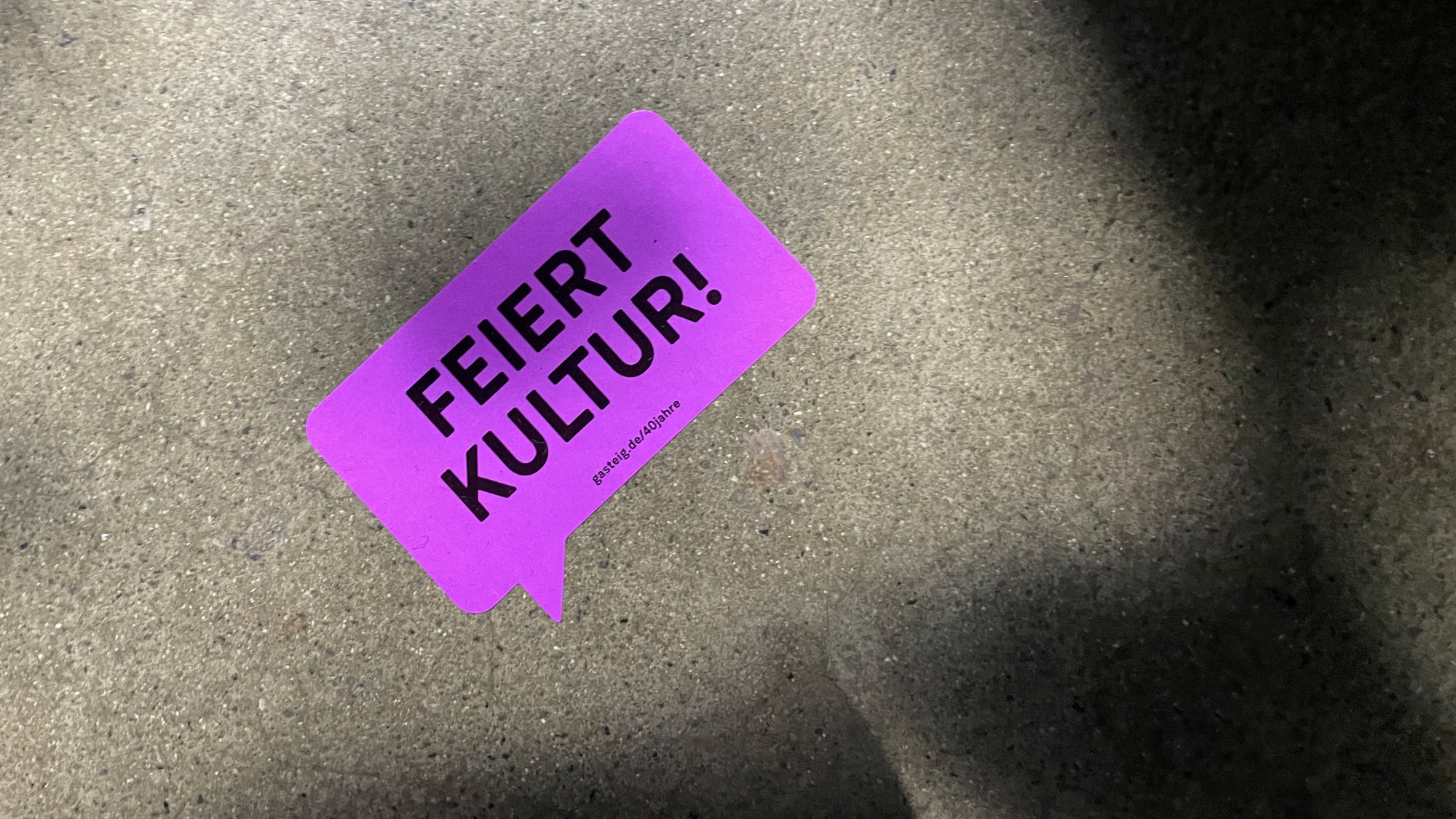 Sticker der Gasteig-Jubiläumskampagne mit dem Claim „Feiert Kultur!“ auf Asphalt – typografisches Kampagnenelement in leuchtendem Pink.