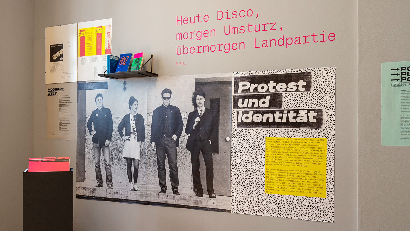 Eine große schwarz-weiße Fotografie mit verschieden großen Plakaten, einem Bücherregal und dem Schriftzug „Heute Disco, morgen Umsturz, übermorgen Landpartie“ an einer Wand in der Ausstellung.