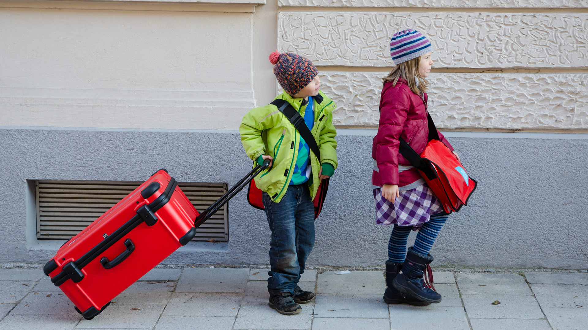 Zwei Kinder auf dem Geweg ziehen den großen roten Koffer hinter sich her.